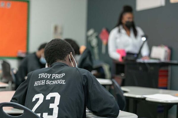 The back of a student sitting at a desk with a shirt that reads "troy prep high school"
